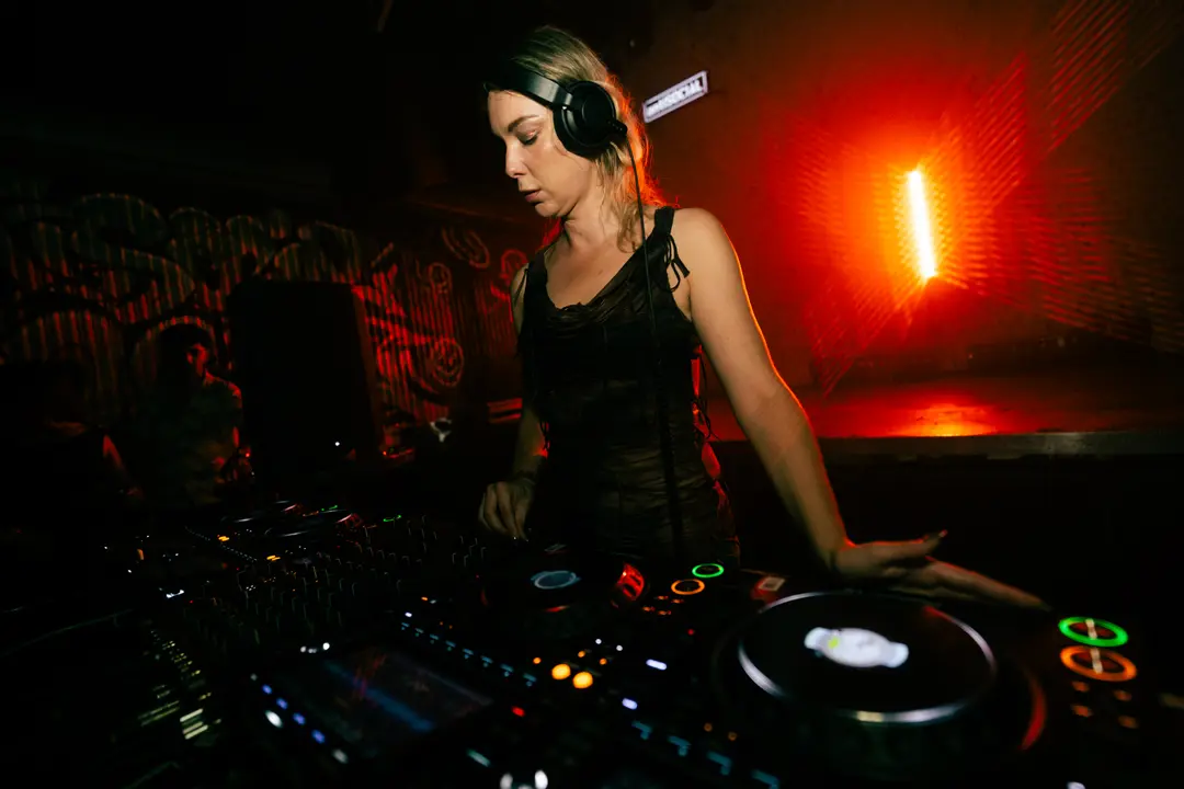 Anastasia Kristensen working the mixer under a red wall light, headphones pushed back on her head
