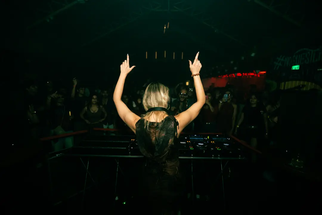 Anastasia Kristensen from behind, both arms raised above the decks, the crowd facing her in the dark